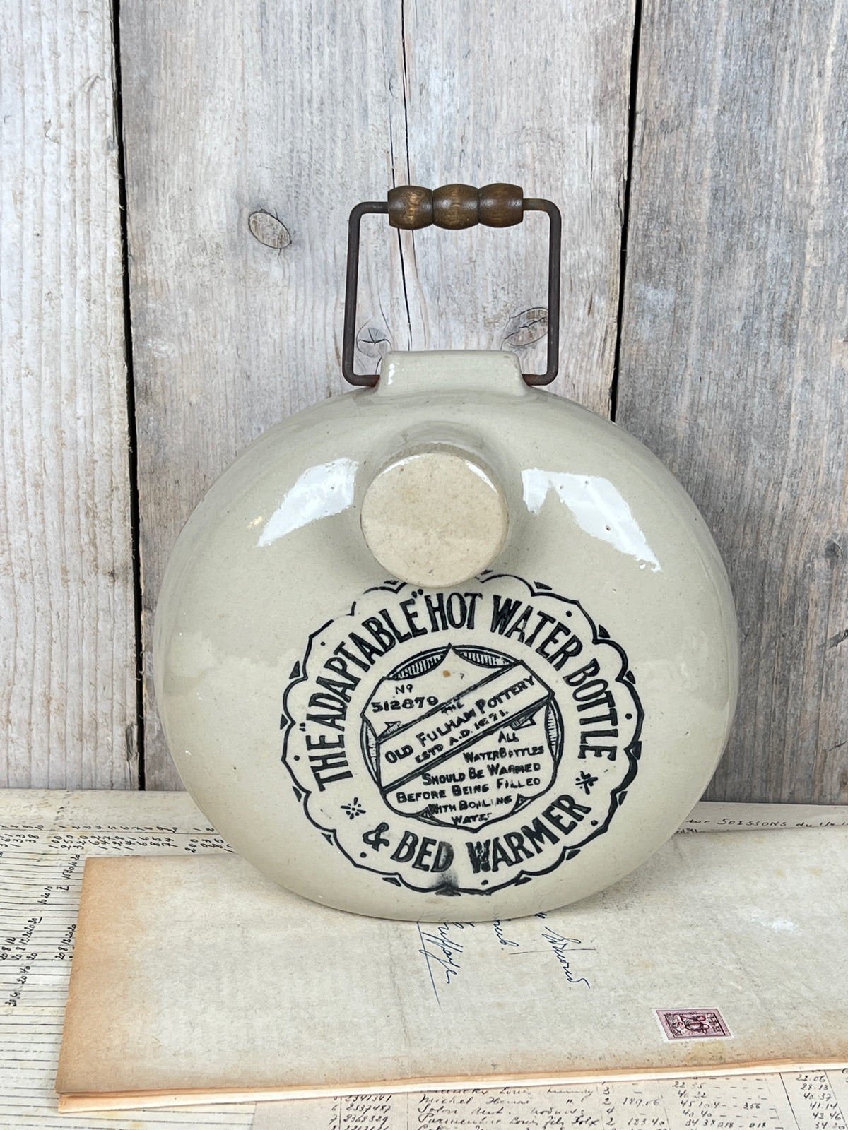 Ancienne bouillotte ronde en grès vernissé beige de la marque anglaise The Old Fulham Pottery avec bouchon et poignée métallique à perles de bois.