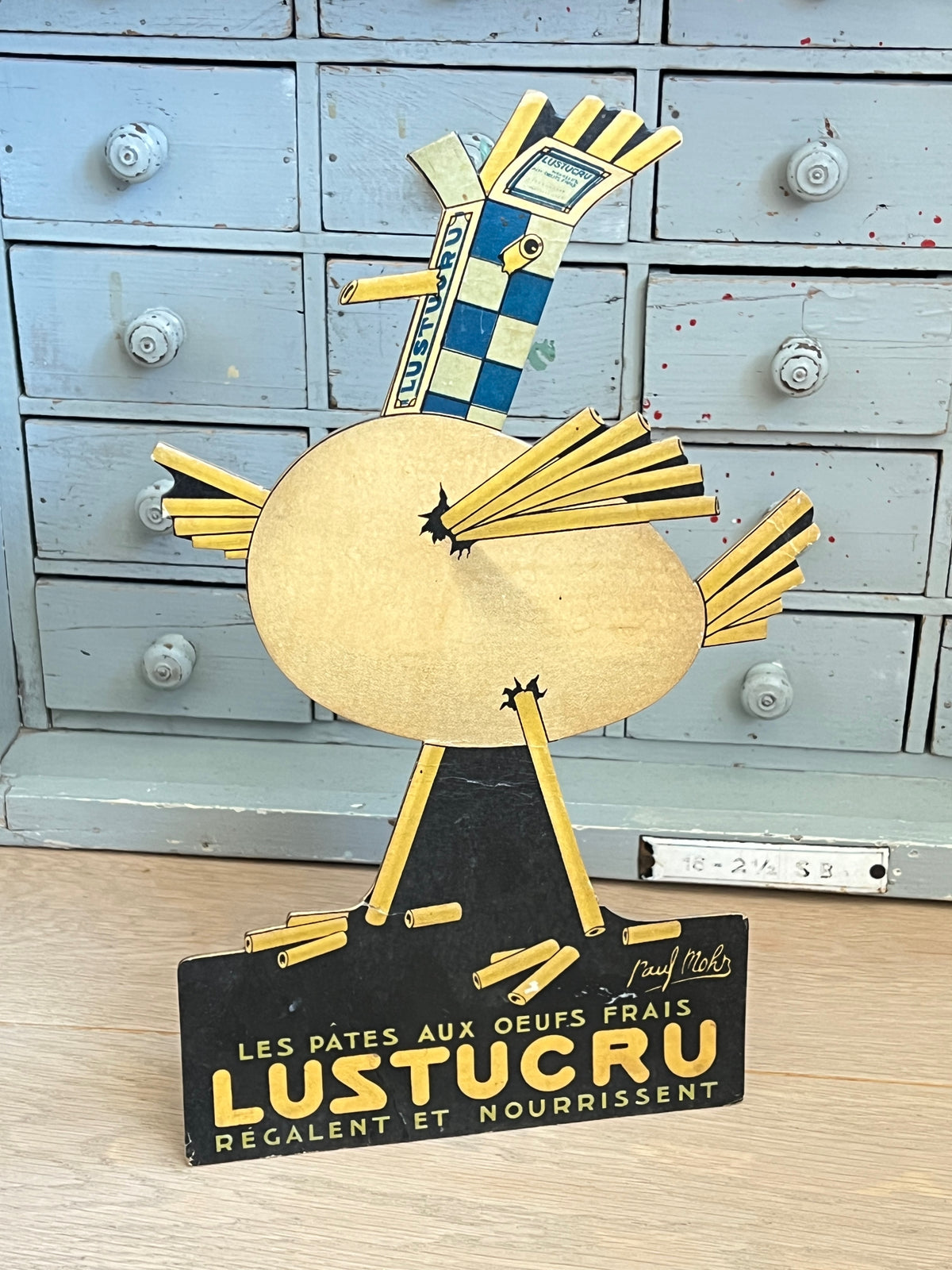 Ancien présentoir publicitaire de comptoir en carton découpé Lustucru par Paul Mohr, forme de poule en pâtes et damier bleu, avec son chevalet au dos.